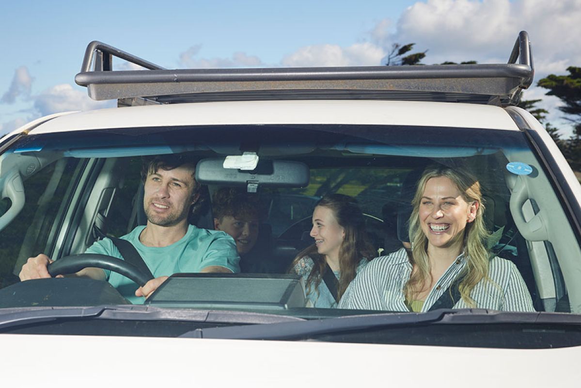 Family in white SUV at coastal location all smiling, with man driving, woman in front passenger seat and boy and girl in back seat.
